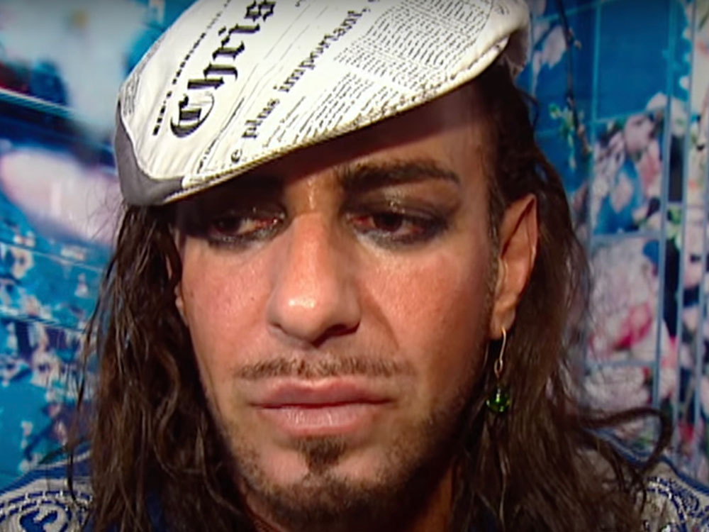 John Galliano has long wavy brown hair and is wearing a pageboy cap with a black and white newsprint design on it. He is seated against a blue floral background, wearing heavy eye makeup and looking down to the left of the frame, frowning.