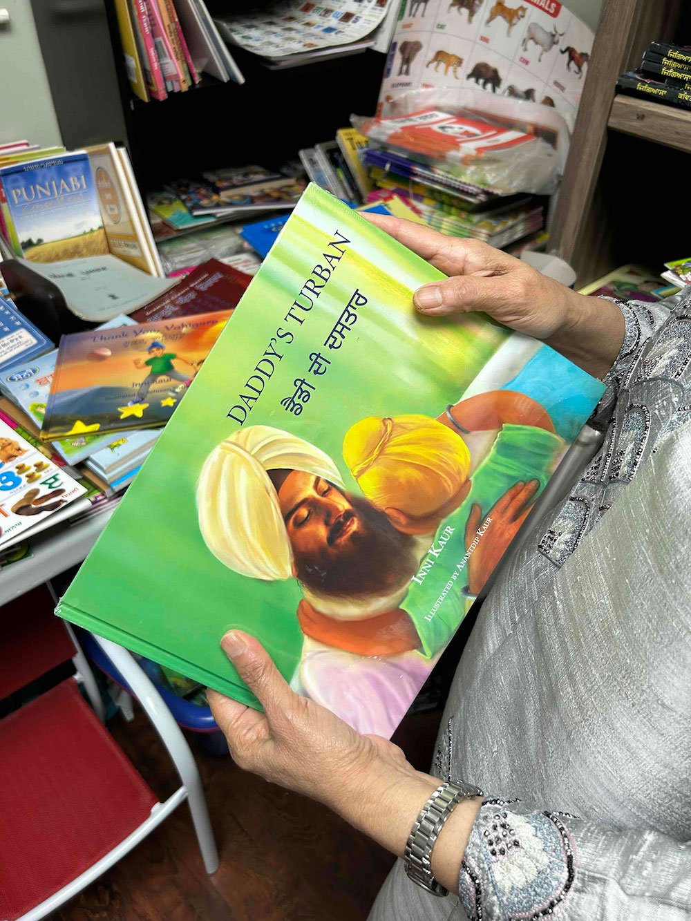 A person wearing a silver suit holds a bright green children’s book titled 'Daddy’s Turban,' featuring an illustration of a Sikh man wearing a yellow turban embracing a child. In the background is the interior of a bookstore full of books.