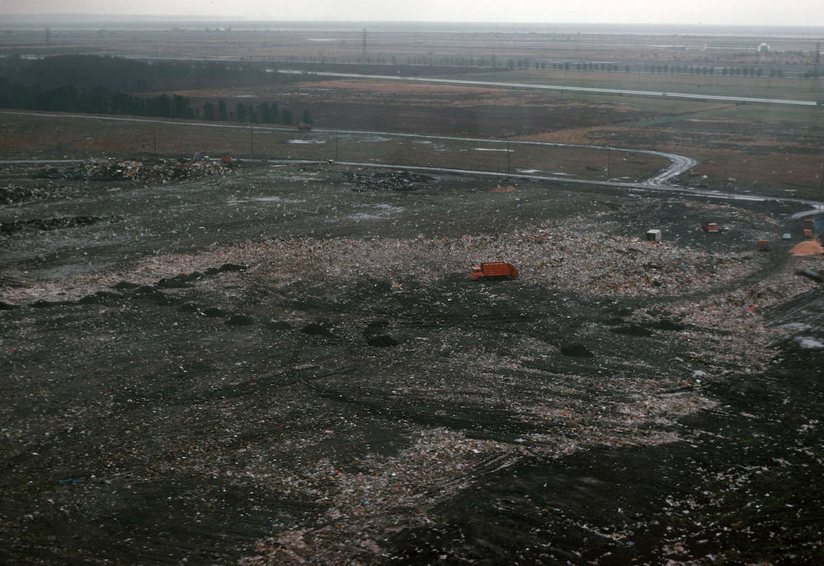 A photograph from 1975 showing a red truck moves among the waste and dirt of a former landfill. In the background is a view of what is likely Richmond.