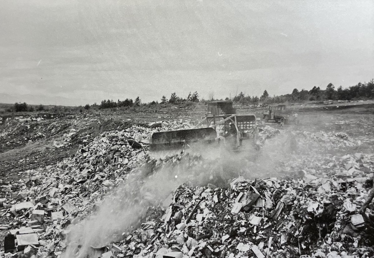A black and white photo shows industrial machines pushing around mounds of garbage at the Kerr Road Dump. Trees surround the site in the background.