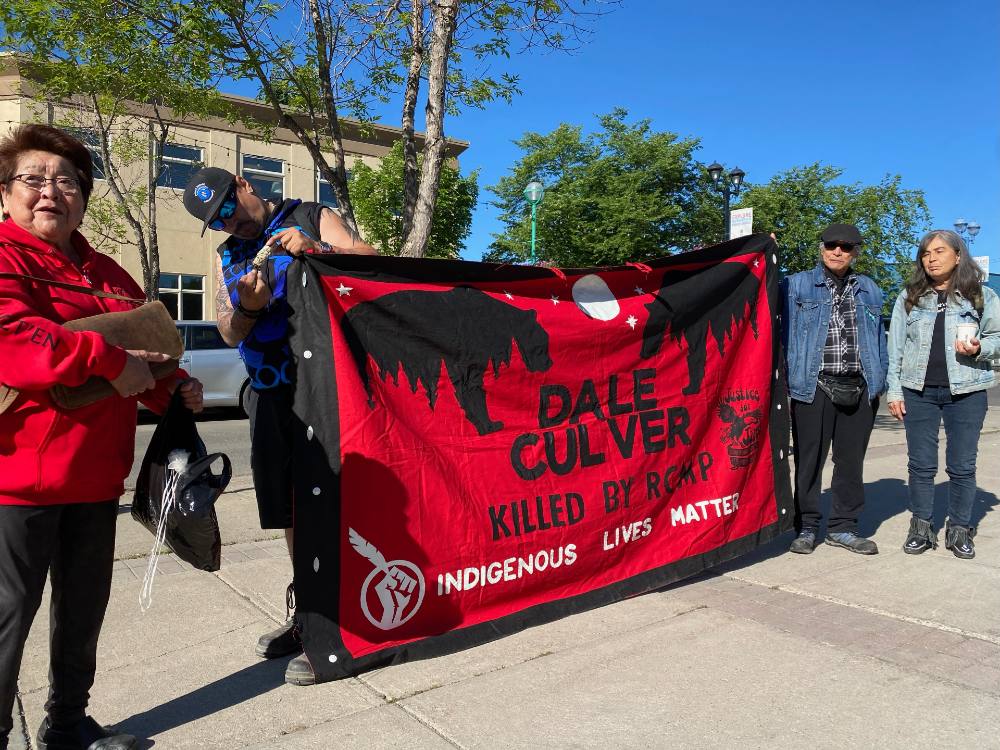 A red and black banner several feet high with the words 'Dale Culver killed by RCMP' and 'Indigenous lives matter' is held up by two people on either side.