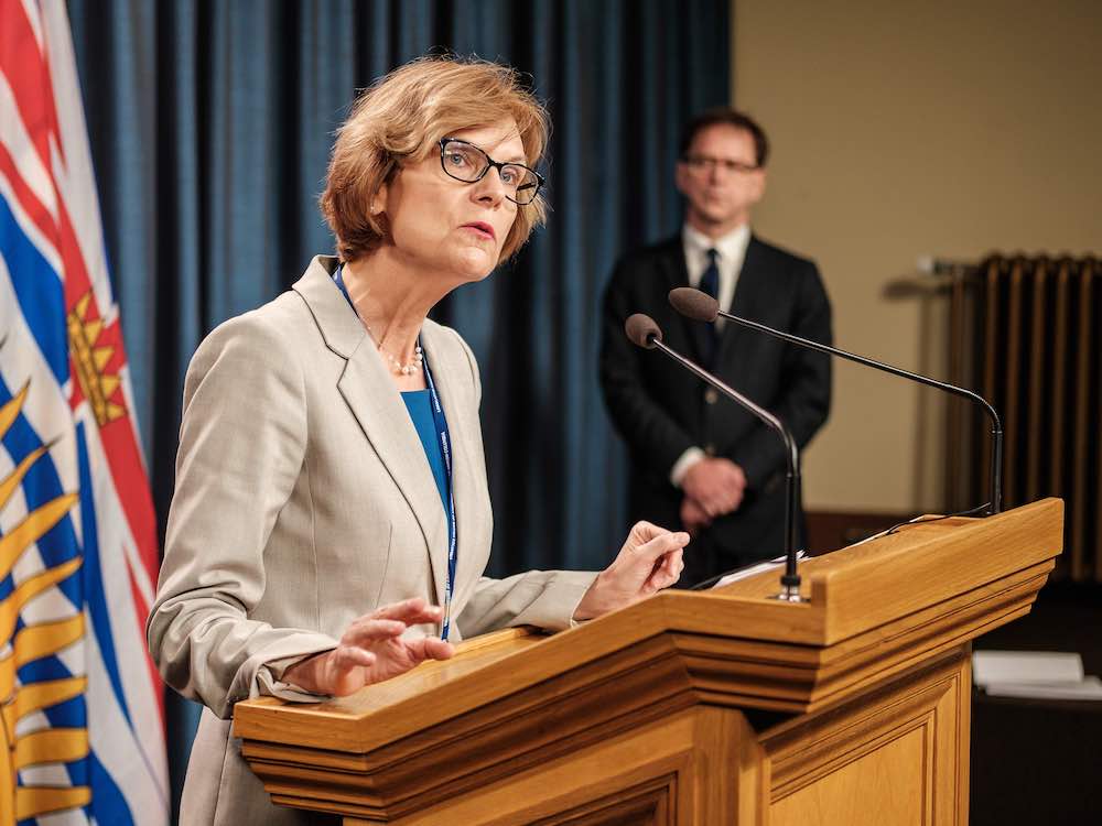Isobel Mackenzie, a middle-aged white woman with a brown bob, stands in front of a wooden podium addressing a press conference. Health Minister Adrian Dix is visible in the background. 
