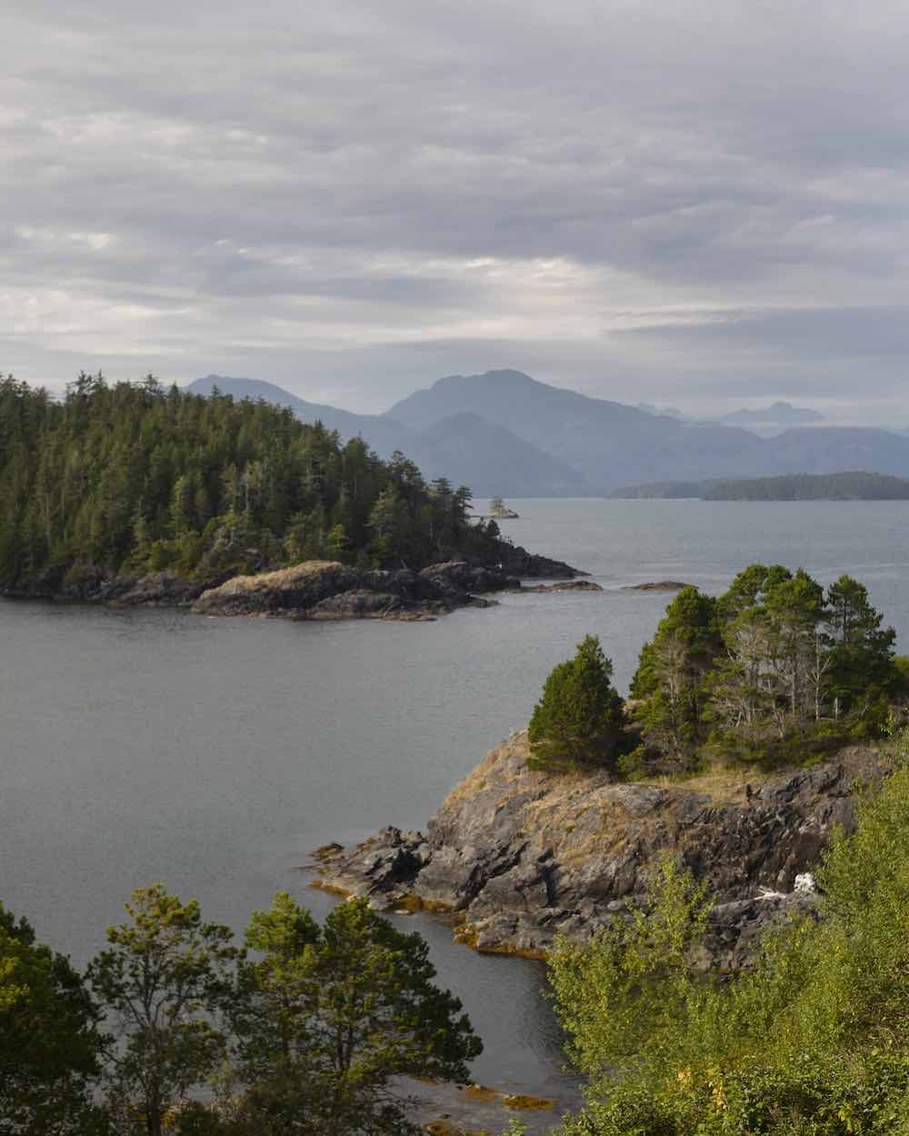 A view of a peaceful bay surrounded by coastal islands covered in coniferous trees. The sky is cloudy and greyish blue. There are blue mountain ranges in the background.