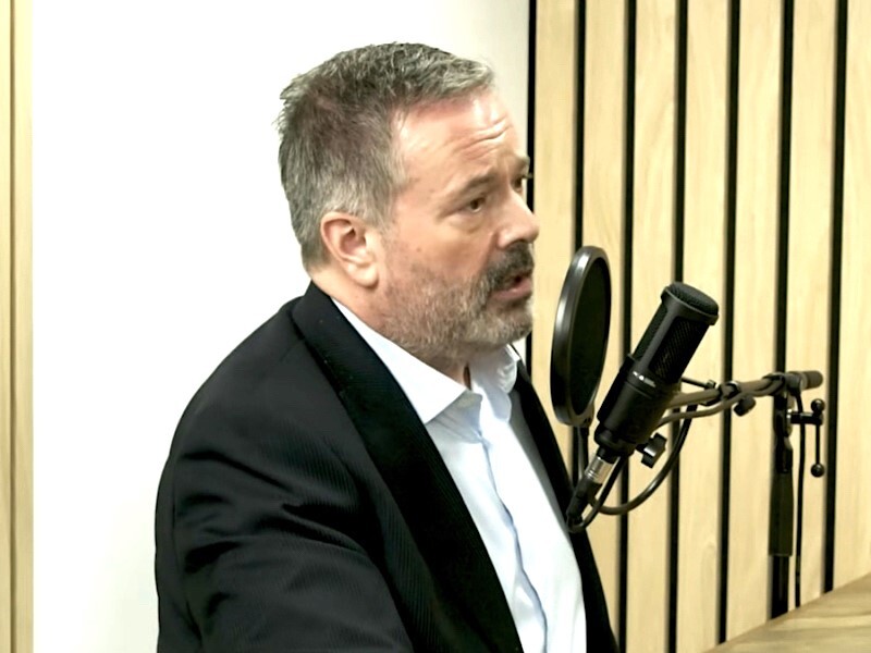 A 55-year-old white man with short greying hair and a scruffy grey beard, wearing a dark blazer and white shirt, speaks into a microphone.