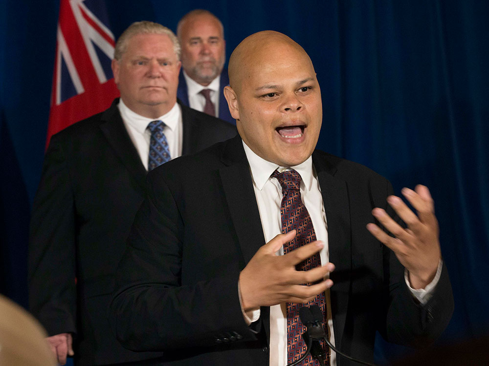 A middle-aged man with medium-dark skin, a bald head and dark suit, white shirt and tie speaks at a press conference. Behind him is a beefy man with similar clothing and slicked back blonde hair.