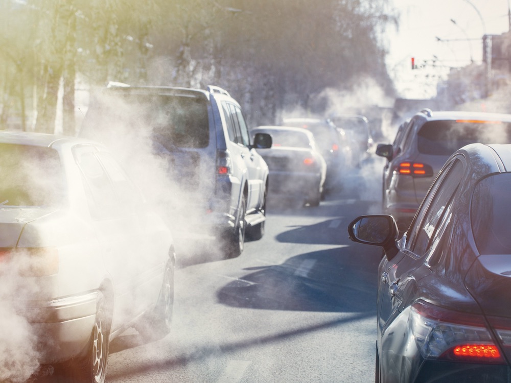Two lanes of cars in high traffic on a cold, sunny winter day are expelling diesel exhaust. The exhaust is visible in the air as white clouds around the cars, which are facing away from the camera.