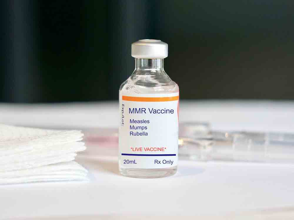 A small clear glass bottle with a silver lid has a white label on it that reads 'MMR vaccine: measles, mumps, rubella. Live vaccine.' The bottle is on a white tabletop with a stack of white compresses to the left.