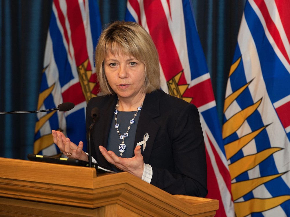 A light-skinned woman with shoulder-length blond hair stands at a podium with B.C. flags behind her. She is in mid-speech with her palms facing upward. She is wearing a black blazer and an unusual necklace made of large white and blue patterned beads.