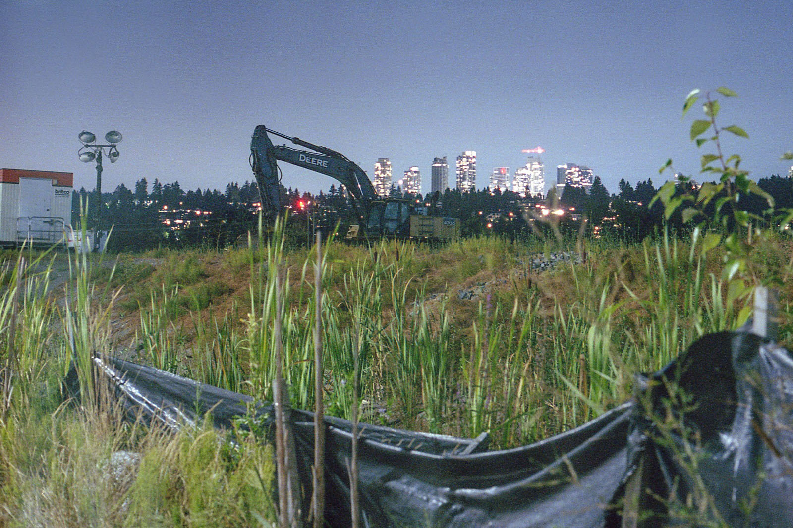 A pile of black debris sits in a tall grass field at night. An excavator is parked in the far centre of the frame. Behind it, a hill with trees is lit with residential lights and a row of highrise buildings in the distance. The sky is dark blue.