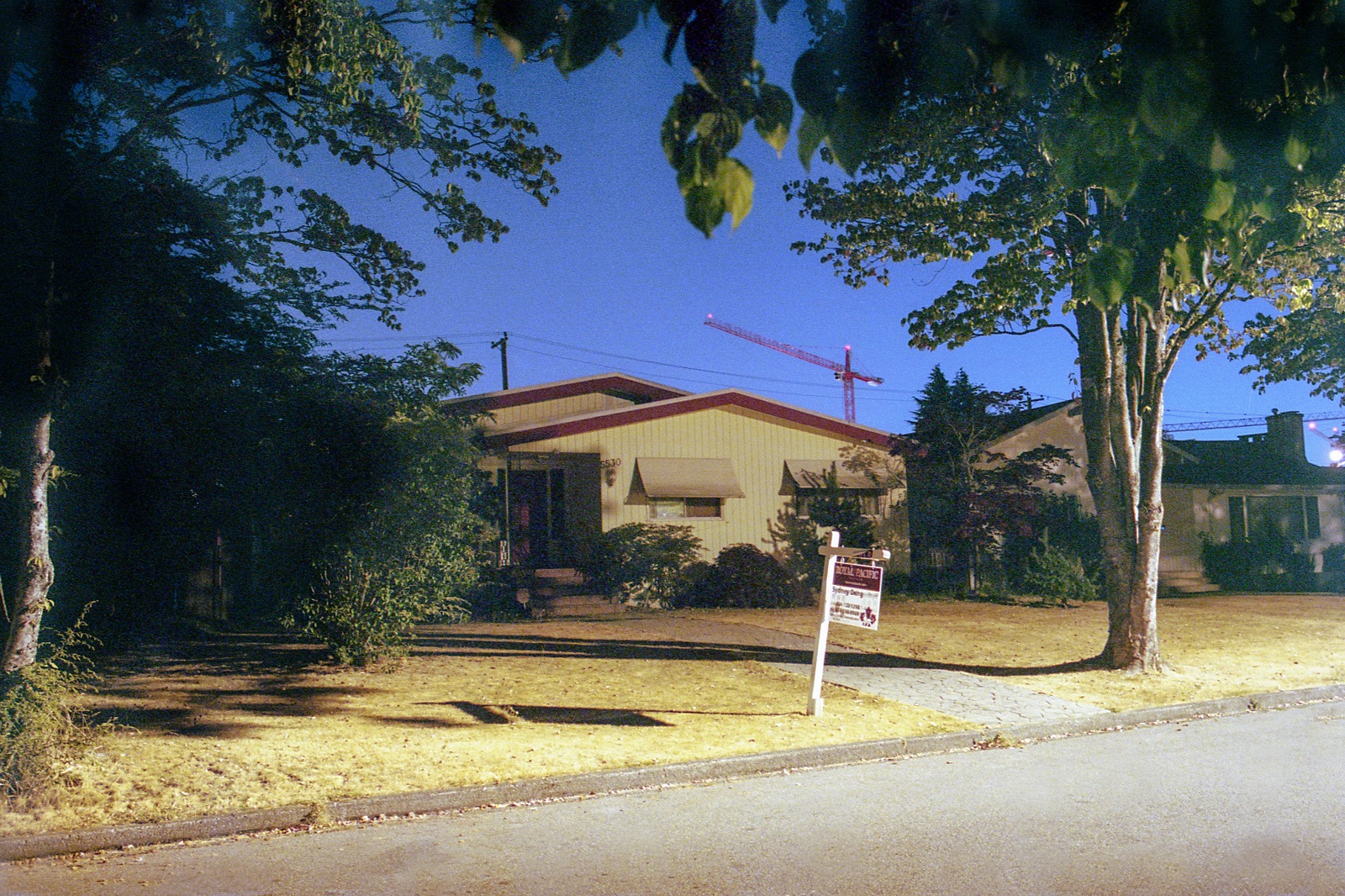 A one-storey beige bungalow lined with shrubbery has a for-sale sign on the front lawn beside a concrete walkway. A blue night sky with construction cranes looms in the background.