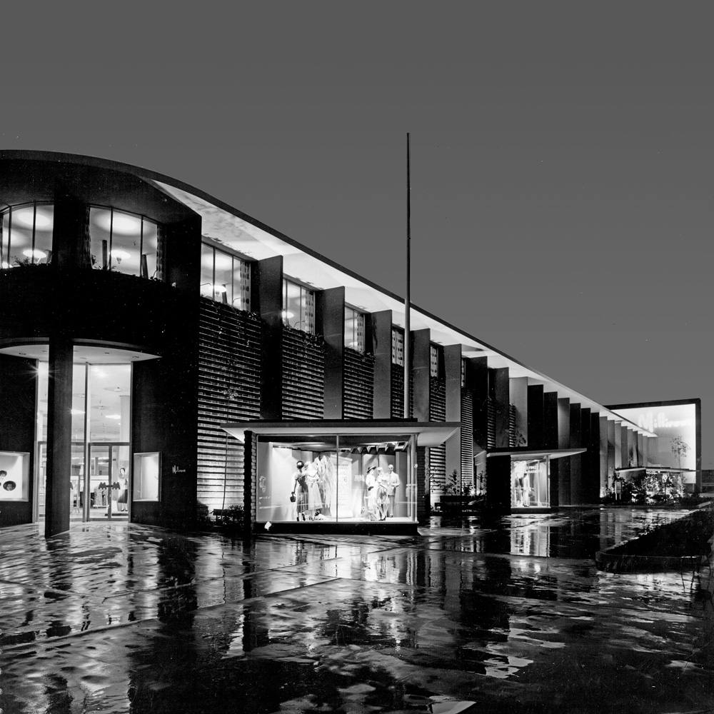 A sepia image of a 1949 department store at night. In the distance is the logo Milliron. There are three enticing window displays with the lights still on and stylish mannequins inside. It just rained and there is a reflection of the store on the wet concrete.
