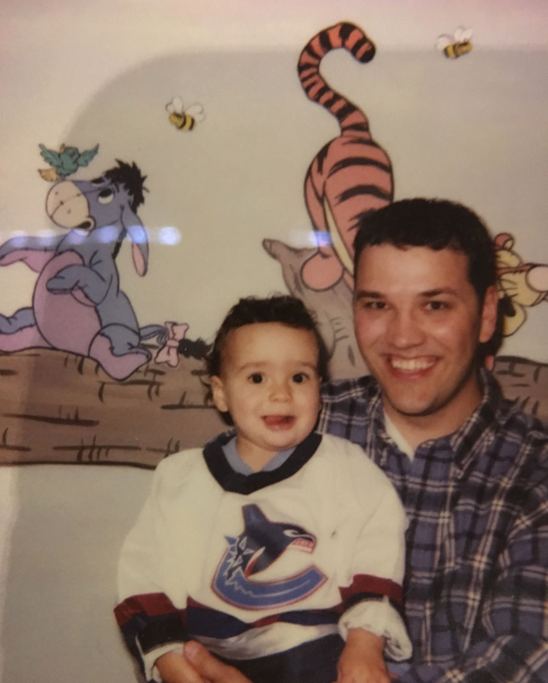 A family photo featuring Josh Kozelj, left, as a toddler. His father is holding him in his arms. Josh is wearing a white Vancouver Canucks hockey jersey. His father is wearing a blue plaid shirt. They are seated against a wall featuring a children’s mural of characters from Winnie the Pooh.