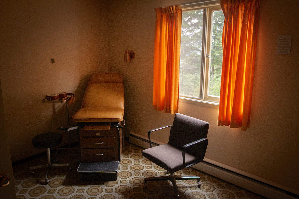 A curtain-framed window lights an empty room, coloured in oranges and beige, with a linoleum patterned floor and a doctor’s examining chair.