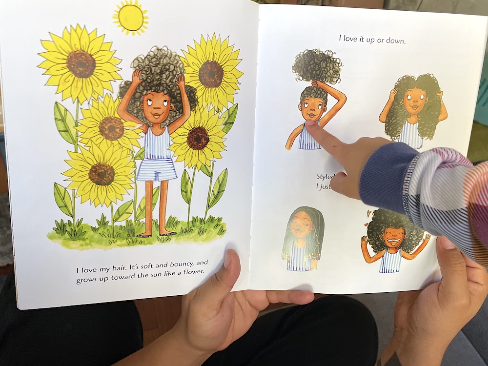 Two hands hold open a children’s book to a two-page spread featuring illustrations of a young Black girl with curly hair. On the left, the girl is standing touching her hair among yellow sunflowers. On the right, the girl is featured in four illustrations demonstrating different hairstyles. To the right of the frame, a child’s hand points to one of the pictures of the girl on the right side of the page.
