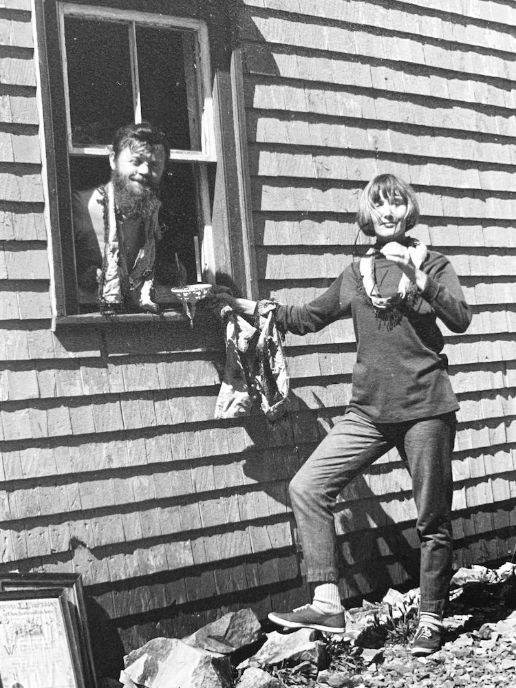 A black and white photo shows Farley Mowat sticking his head out of the window of a house with clapboard siding. Claire Mowat is standing outside.