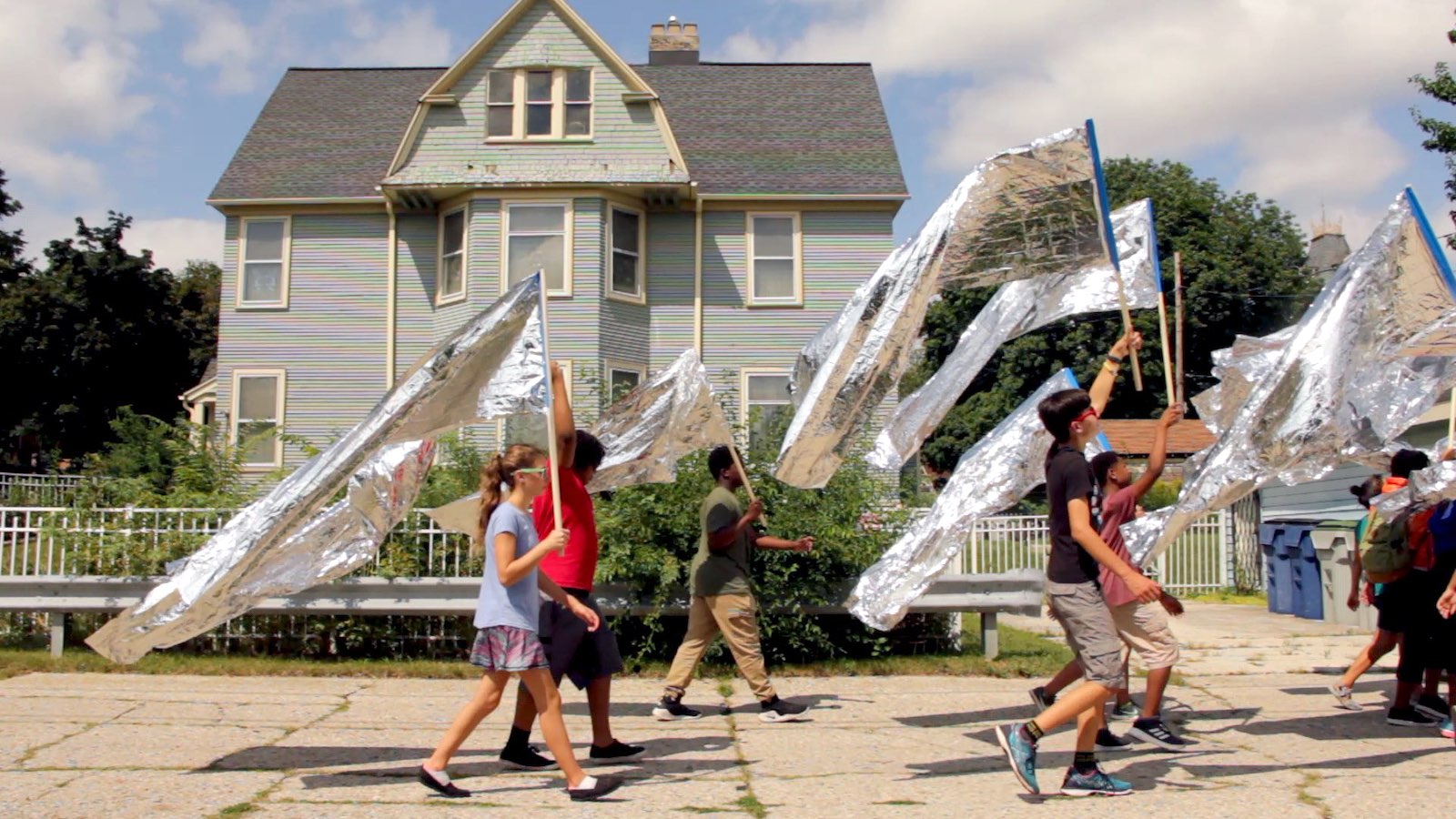 A film video still depicts a group of school-aged children in summer clothes walking down a sunny residential sidewalk, facing the right of the frame. They hold flags on long wooden poles made of silver space blankets. Behind them is an aging, light blue, three-storey house and rows of residential recycling and garbage bins.