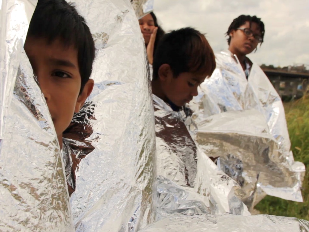 A video still features several young children with dark hair and medium and medium-dark skin wrapped in silver space blankets, their faces partially visible. They are standing in a grassy field.
