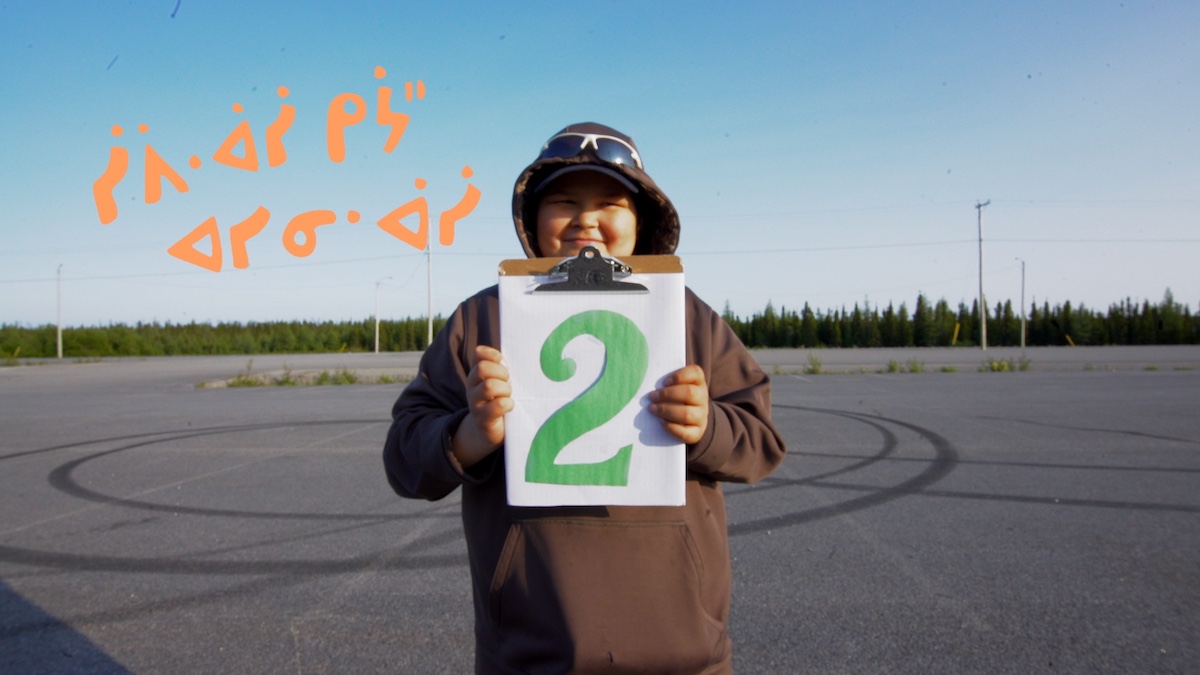 An Indigenous teen in a brown hoodie holds up a clipboard with a green stylized number 2 across the white page. They are standing in an empty parking lot. In the background is a stand of trees and a bright blue sky. On the left of the frame is an orange phrase in an Indigenous language.