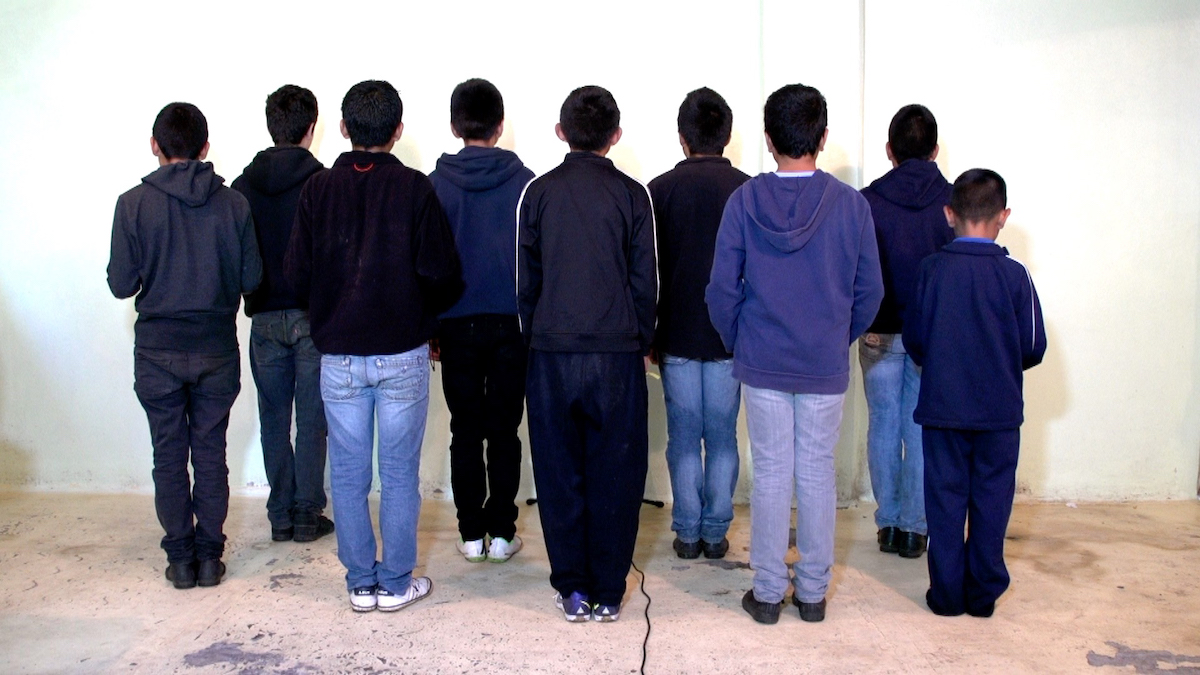 A video still features several children standing on a grey concrete floor with their backs turned to the camera, facing a white wall. They are all wearing dark blue or grey, in jeans or dark athletic pants.