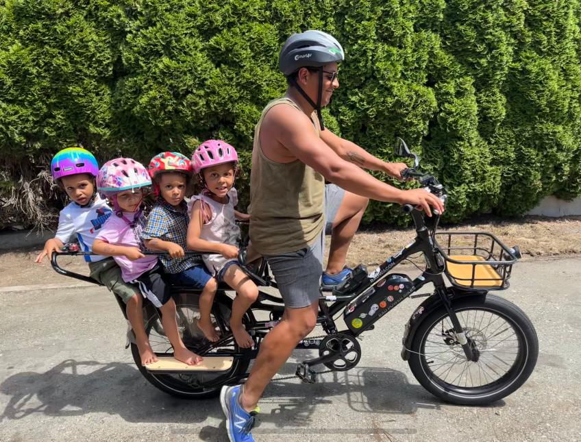 Harrison Mooney helms an e-bike with four young children seated behind him on the cargo bench. They are all wearing colourful helmets. Behind them is a stand of green shrubbery. 