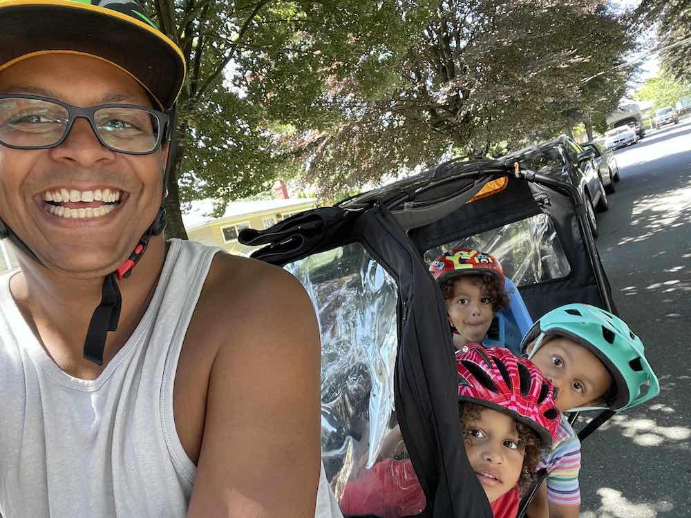 Harrison Mooney is to the left of the frame. He’s smiling and wearing glasses, a tank top and a bike helmet. Behind him in his e-bike’s cargo canopy are three young children in colourful helmets. They’re riding on a quiet residential street on a sunny day.