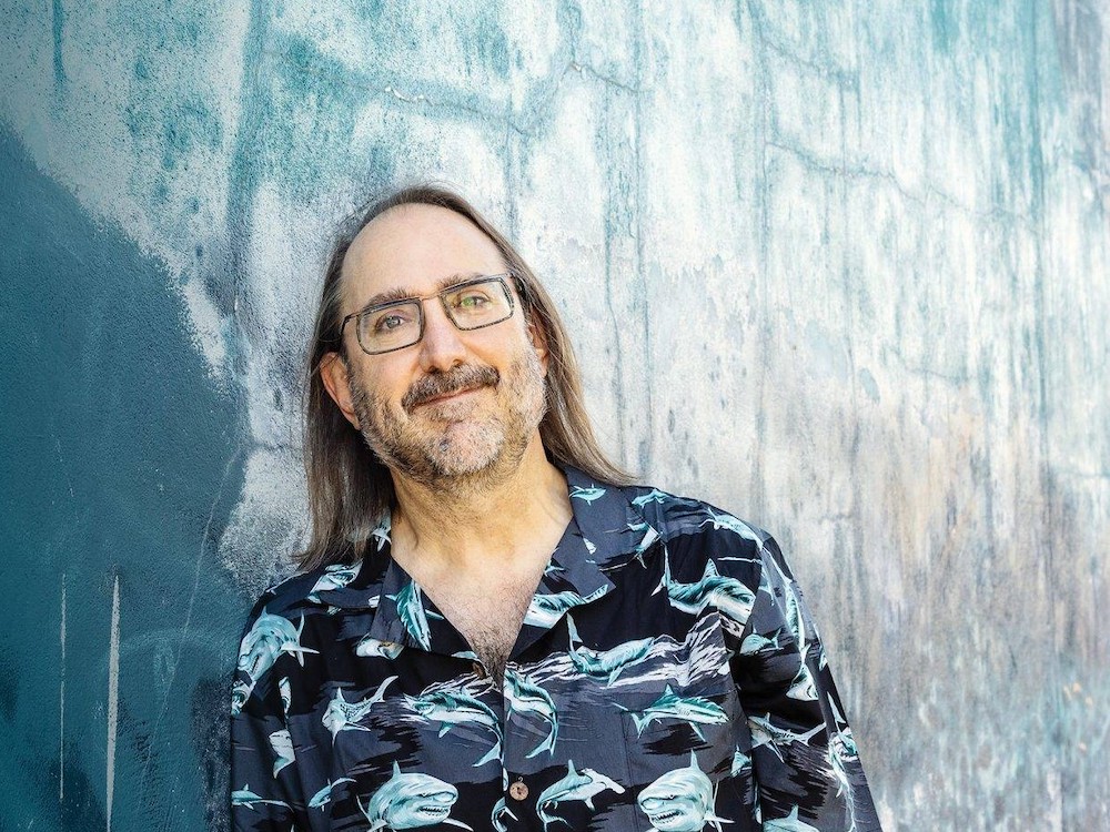 A light-skinned man with long brown hair looks into the camera. He has a salt-and-pepper short-cropped beard and moustache and glasses, and is wearing a shark-print collared shirt.