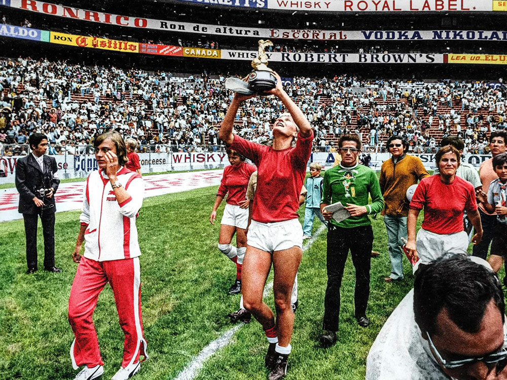 A woman in a red soccer jersey and white shorts holds a gold trophy over her head. She has short blond hair and her light skin is tanned from the sun. She walks across the green pitch of a soccer field inside a crowded stadium. Teammates in similar uniforms and other personnel in casual streetwear from the early 1970s walk near her.