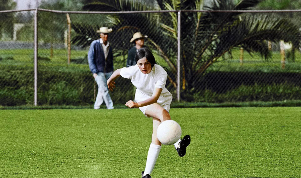 A woman in a white soccer uniform kicks a white soccer ball on a green pitch. She has long dark hair and light skin. Behind her are two men in light-coloured, wide-brimmed cowboy hats.