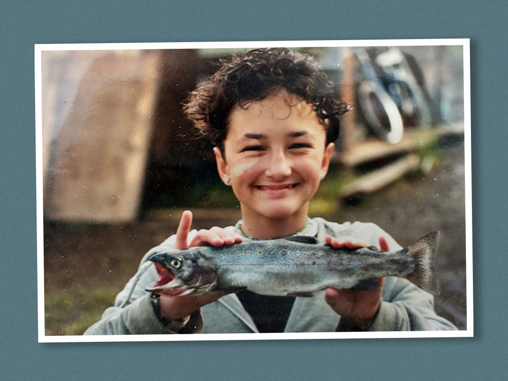 A child with medium-light skin tone and curly dark hair poses with a small fish, smiling. The photo is older, from the 1990s, with a bit of water damage.