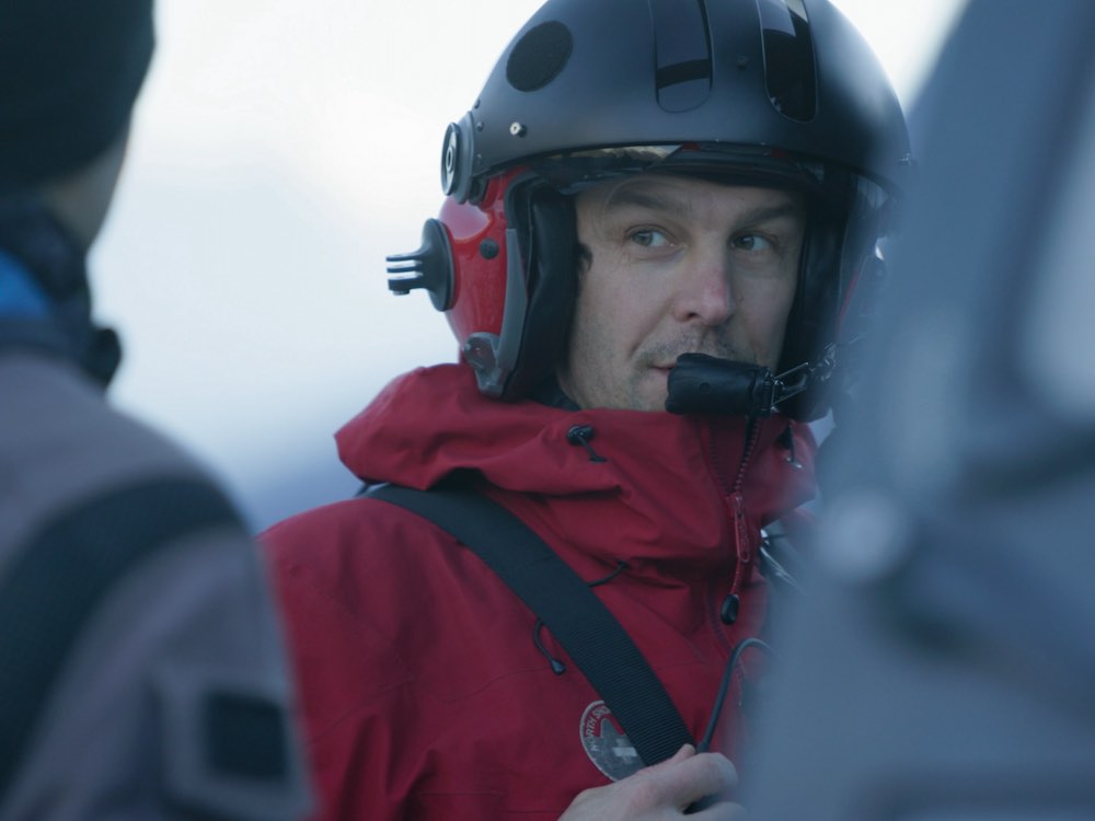 Mike Danks wears a black helicopter helmet and headset with red trim. He has short dark hair, light skin and is looking towards the left of the frame. He wears a red North Shore Rescue team jacket. In the foreground is the soft-focus door of a helicopter.