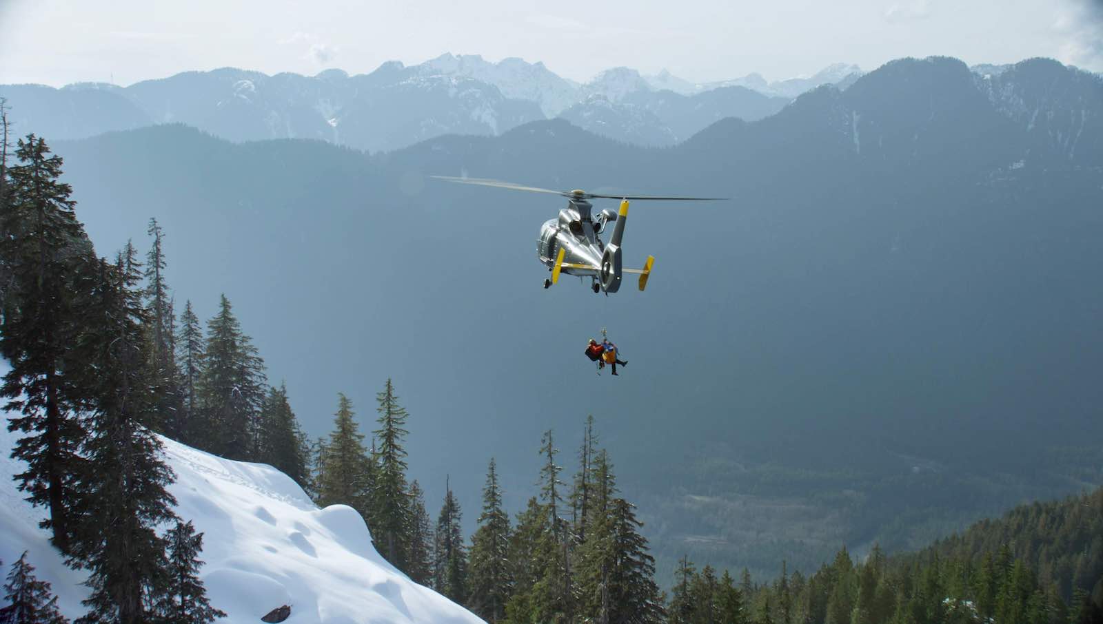 A silver helicopter flies with a longline on which a North Shore Rescue team member in a helmet and red jacket holds an injured person in a stretcher. They are flying against an expanse of green mountains and away from a snowy hill.