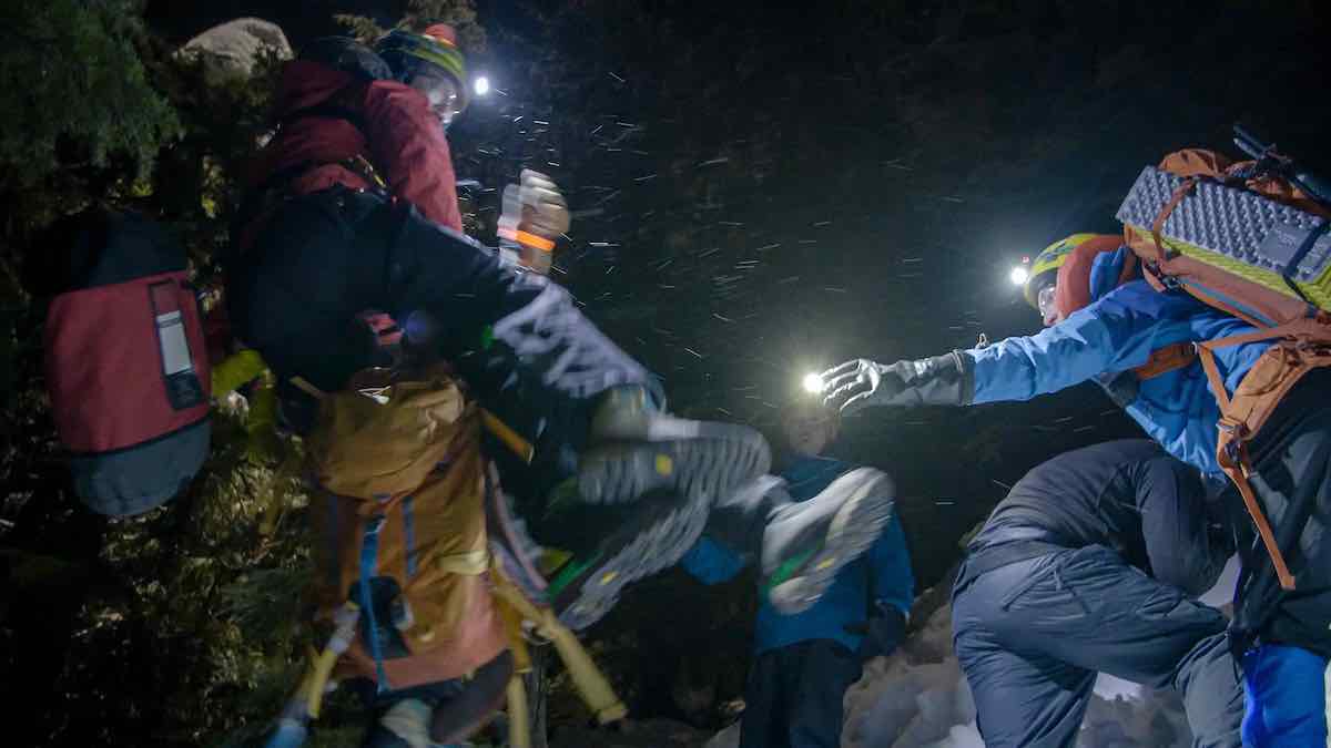 The camera looks up from the ground to a black night sky where snow or rain is falling as a person in a yellow helmet and blue jacket, right, reaches towards a person in another helmet and red jacket, left, who is in the midst of helping hoist an injured person to safety on a helicopter longline.
