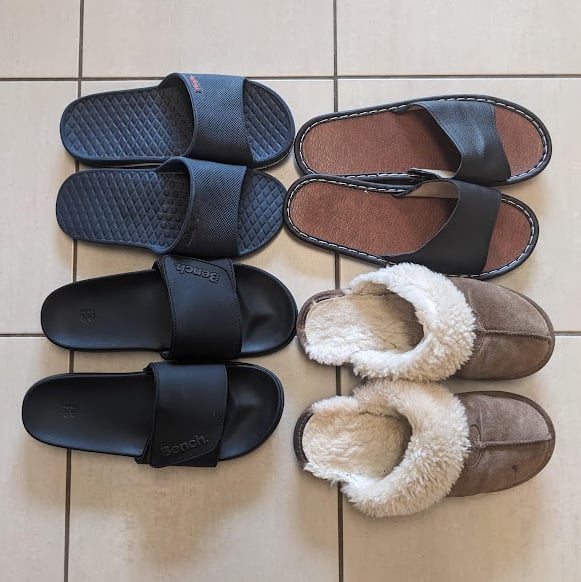 Four pairs of slip-on brown or black sandals or slippers stand on a beige tiled floor facing the right of the frame.