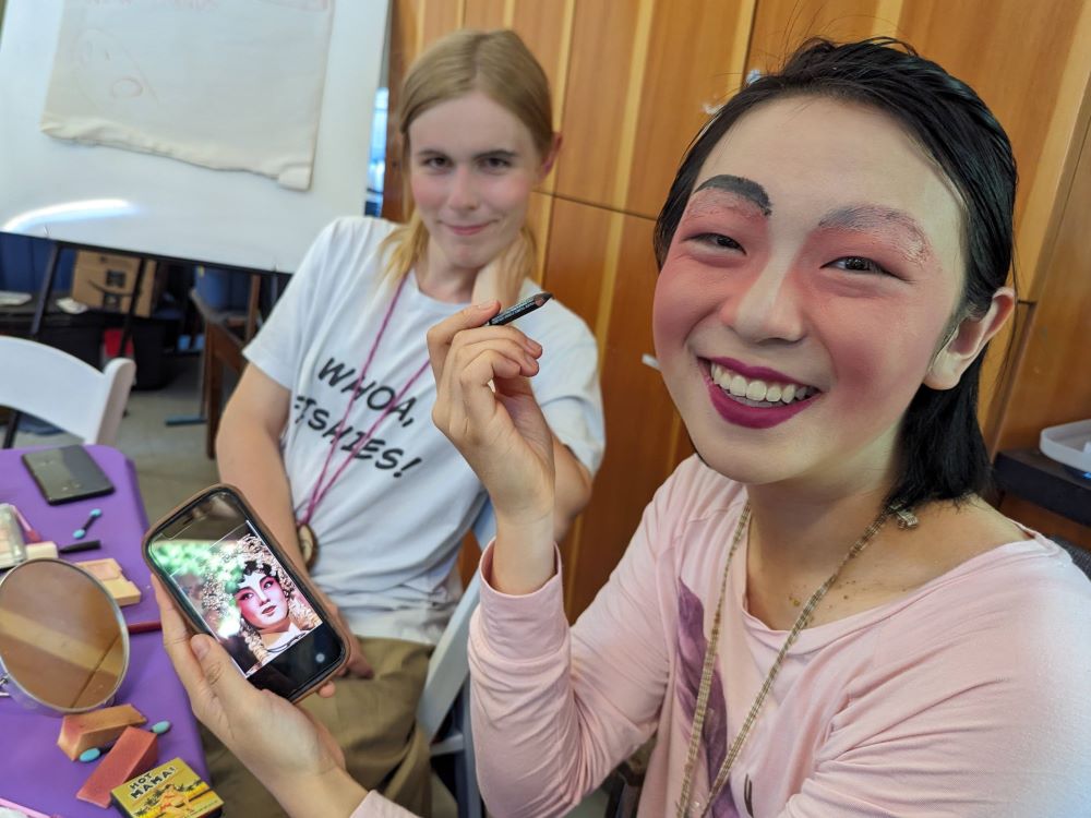 Two young people with light skin tone sit side by-side at a table. The person on the right is applying makeup, using a photo on their smartphone as inspiration.