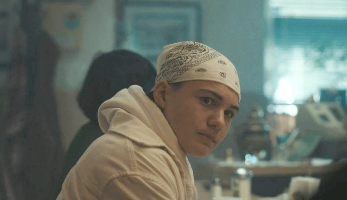 A young teenage boy in a white hoodie with a white bandana on his head looks into the middle distance. He has light skin and dark eyes. Behind him in soft focus is the interior of a diner.