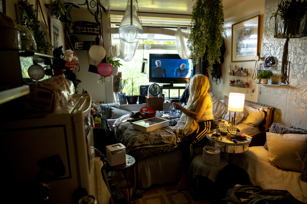 A woman with long bleached-blond hair covering her face sits on a bed in a white tank top and black track pants. She’s in a small, crowded one-room apartment. A television is mounted high on the wall behind her and a large plant with long green vines hangs from a pot from the ceiling.