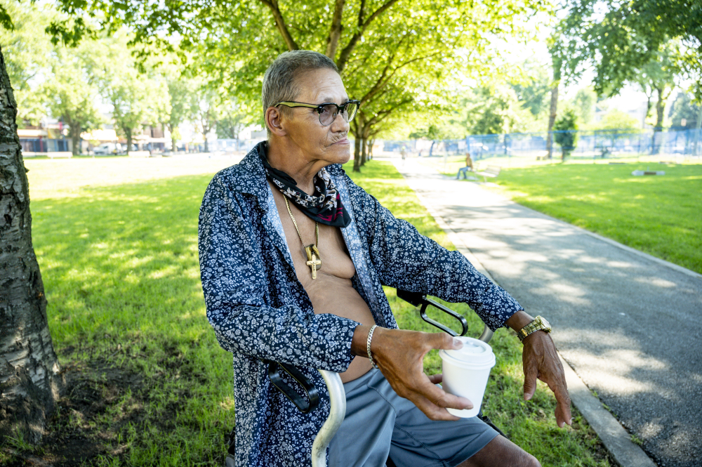 A man with short dark hair in a buzz cut, medium skin and glasses sits on a walker in a sunny Downtown Eastside park near a concrete walkway. His blue button-down floral shirt is open to reveal a bare chest and a necklace featuring a large pendant of a cross. He holds a white paper coffee cup in one hand.