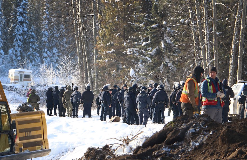 RCMP and land defenders are clustered together in a snowy area. Trees are visible in the background, as is a camper, and some construction equipment.