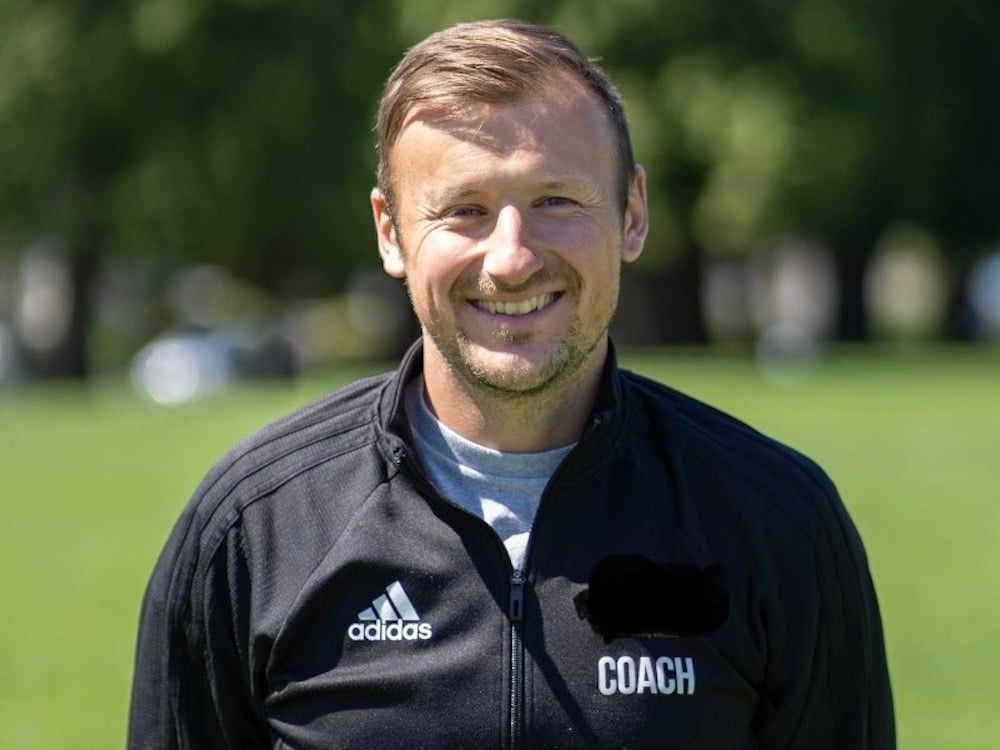 A man wearing a jacket that reads “Coach” stands on a soccer field. He smiles at the camera.