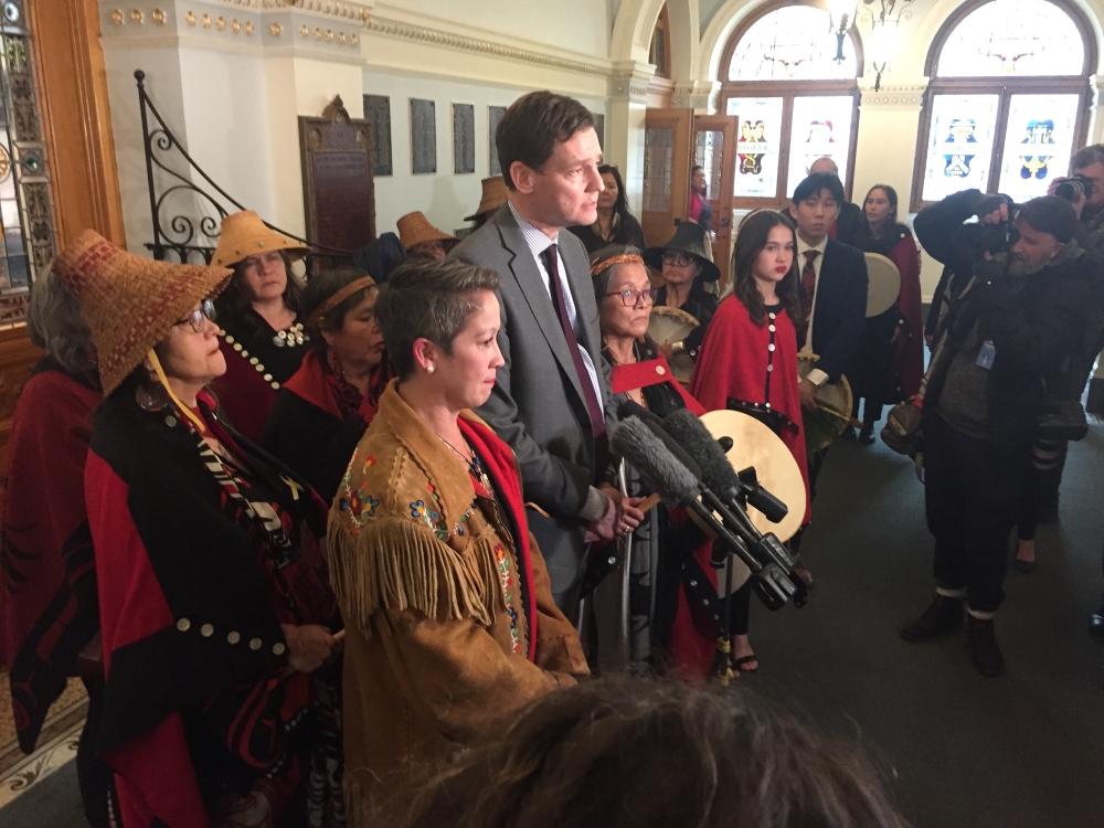Melanie Mark, a woman with short brown hair wearing a fringed leather coat with beadwork, stands before a microphone. Next to her is B.C. Premier David Eby, tall in a grey suit and tie. Behind Mark stands two older women wearing traditional Indigenous woven hats.