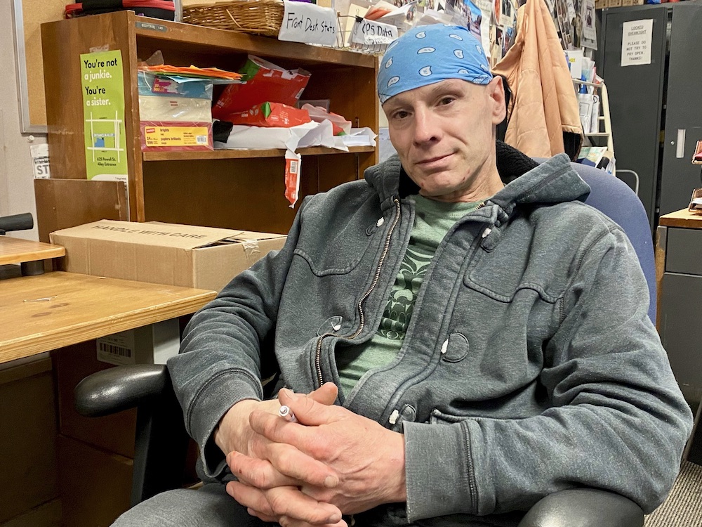 Dennis Hunter sits in a chair, smiling at the camera and wearing a grey jacket and blue bandana tied around his head.