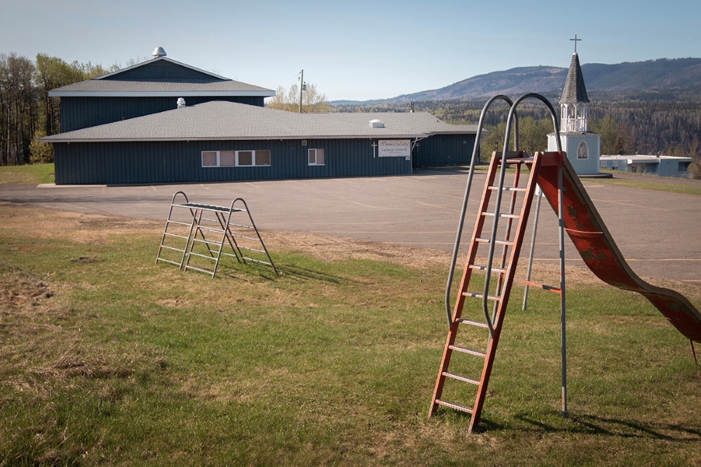 A couple old metal pieces of playground equipment on the lawn in front of a blue building. It’s a sunny day, and there are hills in the background.