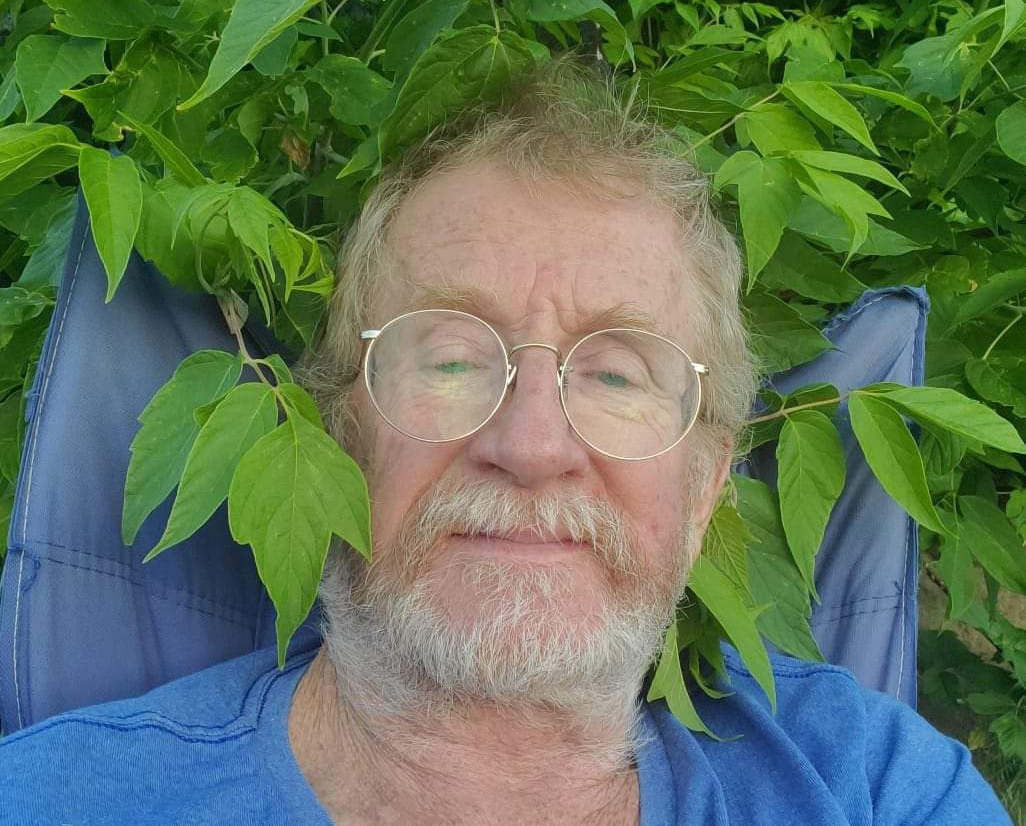 A close-up of a man’s face, with greying hair and beard, and wearing glasses. He sits in a blue lawn chair surrounded by green leaves.