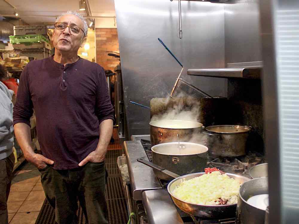A man with short white hair, glasses and a burgundy Henley stands on the left of the frame with his hands in the pockets of his brown corduroys. He is standing in a restaurant kitchen. Beside him, several stainless steel pots simmer on a gas range. In the foreground is a pan piled high with diced onions.