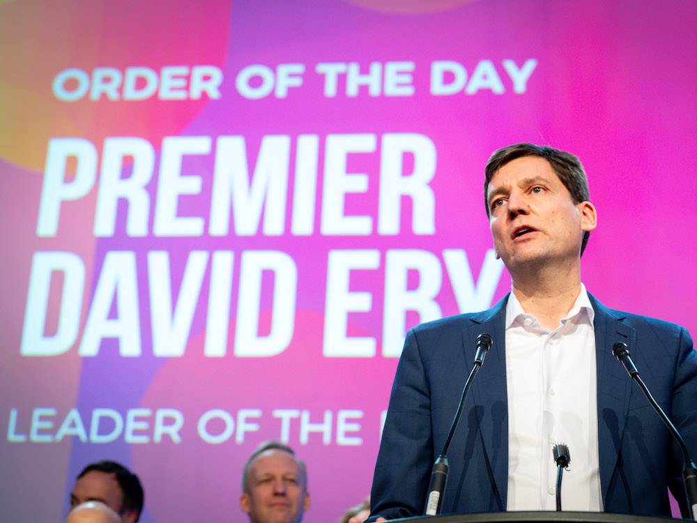 A white man with dark hair, wearing a white shirt and blue jacket, stands at a podium. Behind him on a purple-lighted screen, a sign says 'Premier David Eby.'