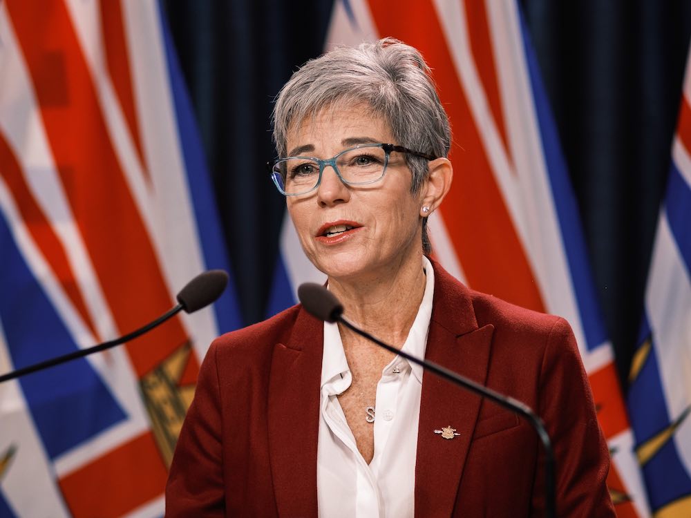 A 60-ish stylish white woman, with short grey hair and light blue framed glasses, stands at a podium, wearing a white shirt and burgundy jacket.