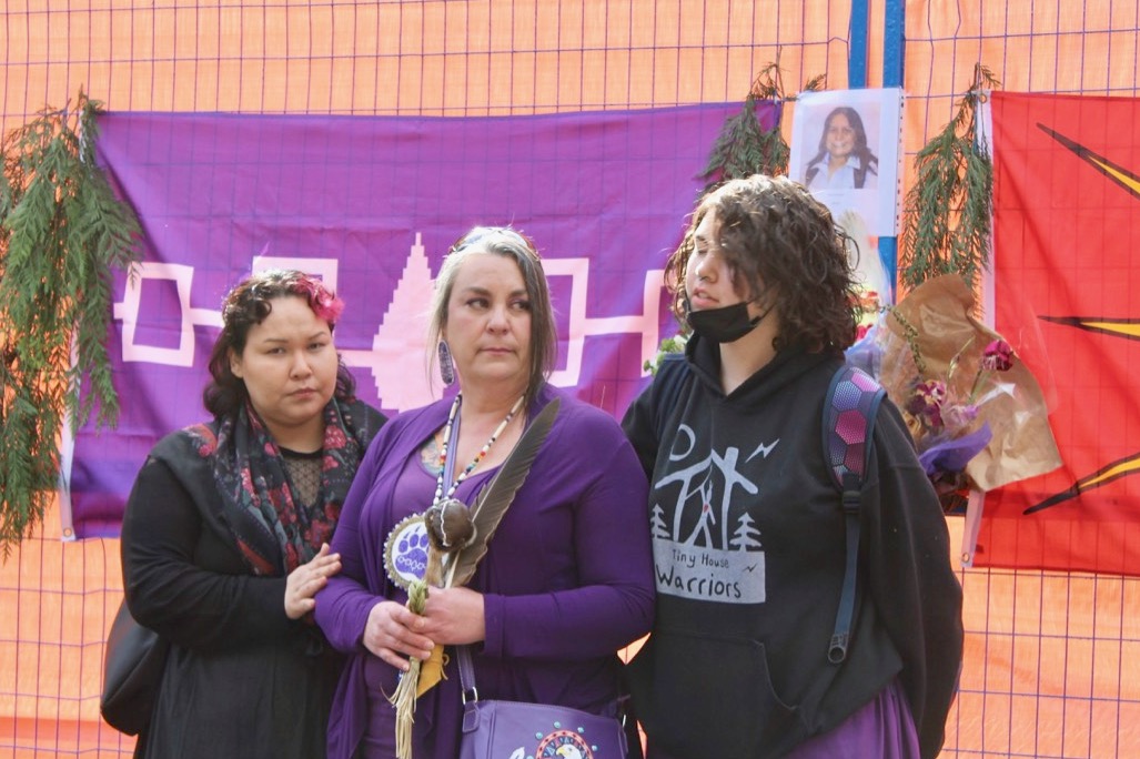 Three people stand in front of a purple banner with a photo of Mary Ann Garlow affixed to the top right corner. Misty Fredericks, in the centre, is wearing purple and is holding a feather and other items.