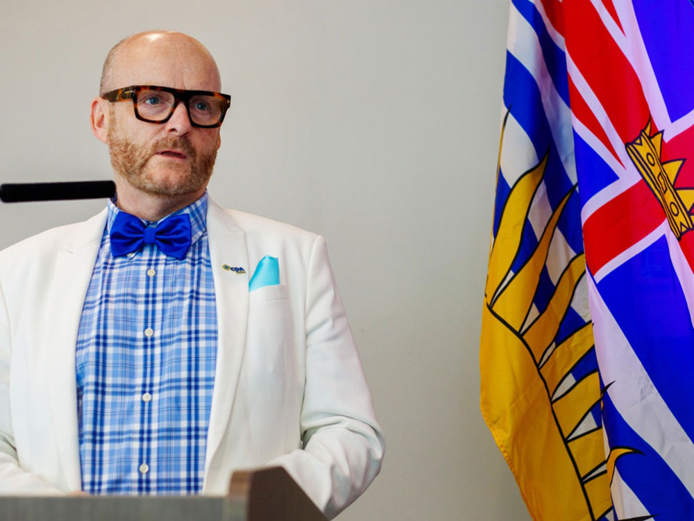 A bald man with thick-framed glasses stands at a podium wearing a white blazer and blue bowtie. Next to him is a flag of British Columbia.