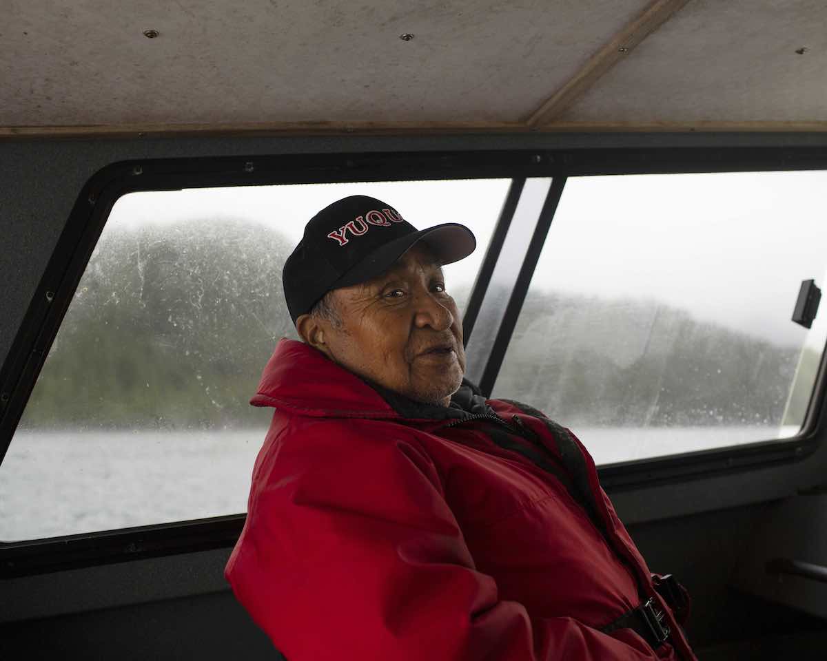 An Indigenous man with medium skin tone wears a black ball cap and a red hoodie. He is seated inside a dimly lit fishing boat. Outside the window behind him is a stand of trees; the sky and water are grey.