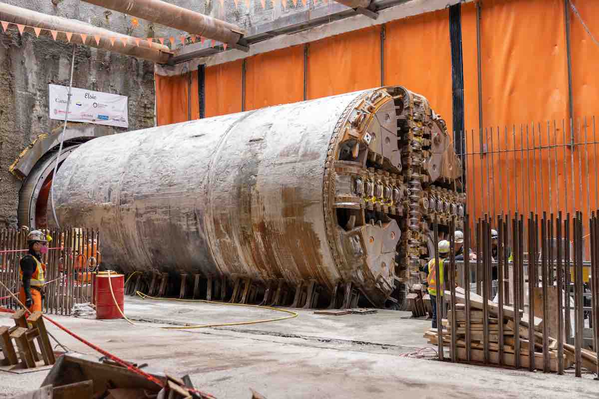 A large cylindrical machine sits in a construction site against a wall covered in orange tarp. In the foreground are construction workers in hard hats and orange and yellow vests.